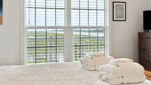 Ein Schlafzimmer mit großem Fenster, einem Bett mit weißer Bettwäsche, einem Holzschrank und einem gerahmten Bild an der Wand.
