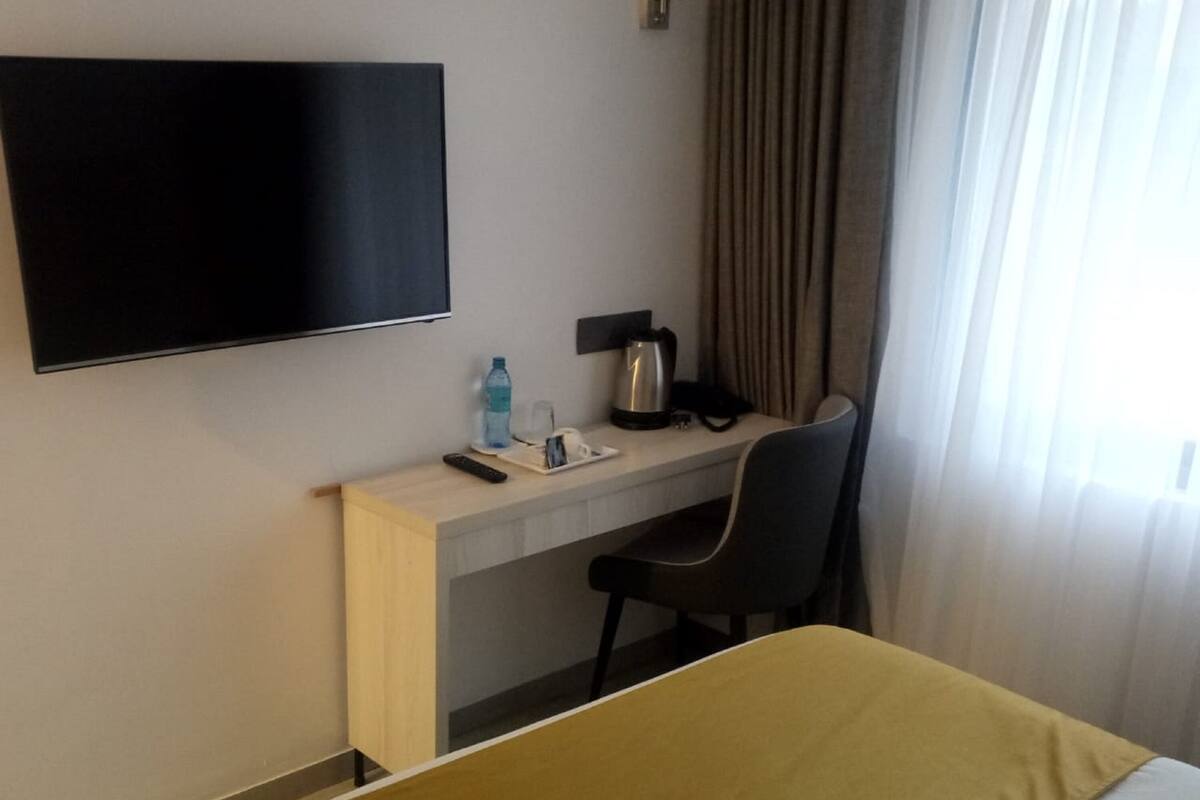 Quarto de hotel com cama, escrivaninha e cadeira, televisão, cafeteira e garrafa de água.