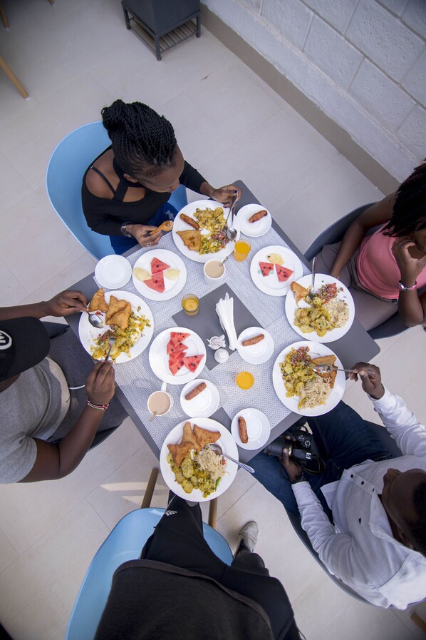 A group of people dining around a table with various dishes, including watermelon slices and a camera on the table. - Symphony Hotel (Naivasha)