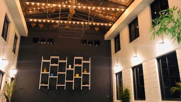 Un interior moderno con techo alto, conductos expuestos y luces colgantes. Varias mesas y sillas dispuestas para comer o socializar.