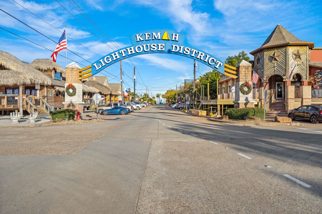 A street view of KEMAH Lighthouse District with thatched-roof buildings, parked cars, and a clear sky.