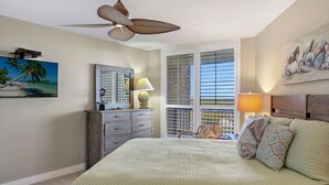 Ein Schlafzimmer mit einem Bett, einem Deckenventilator, einem Schminktisch, einem Stuhl, einem Fenster mit Jalousien und einem Bild an der Wand.
