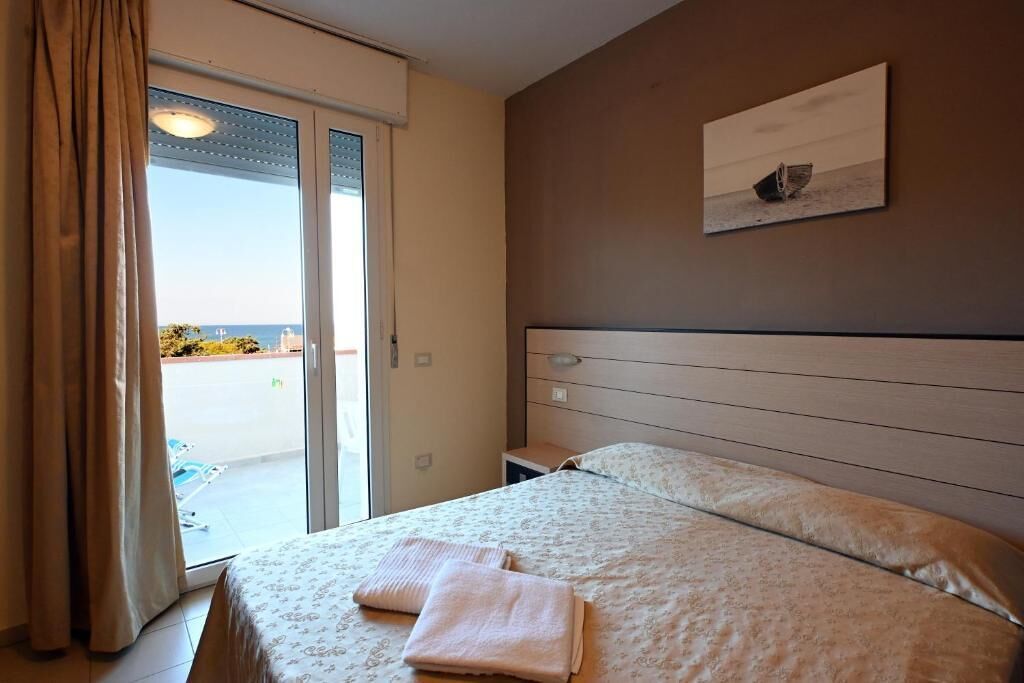 Una habitación de hotel con una cama, una mesita de noche, un cuadro en la pared y vistas a través de la puerta.