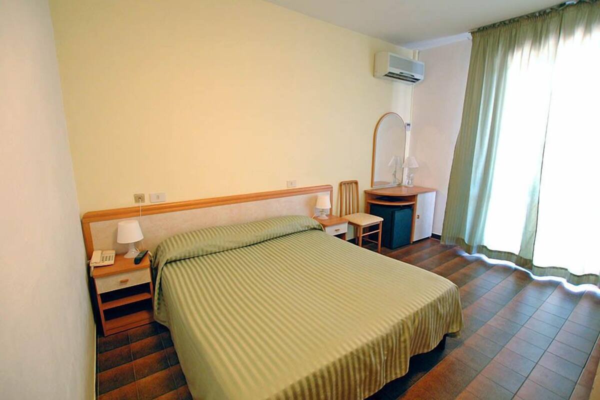 A hotel room with a bed, a desk, a chair, a lamp, a nightstand, and a window with curtains.