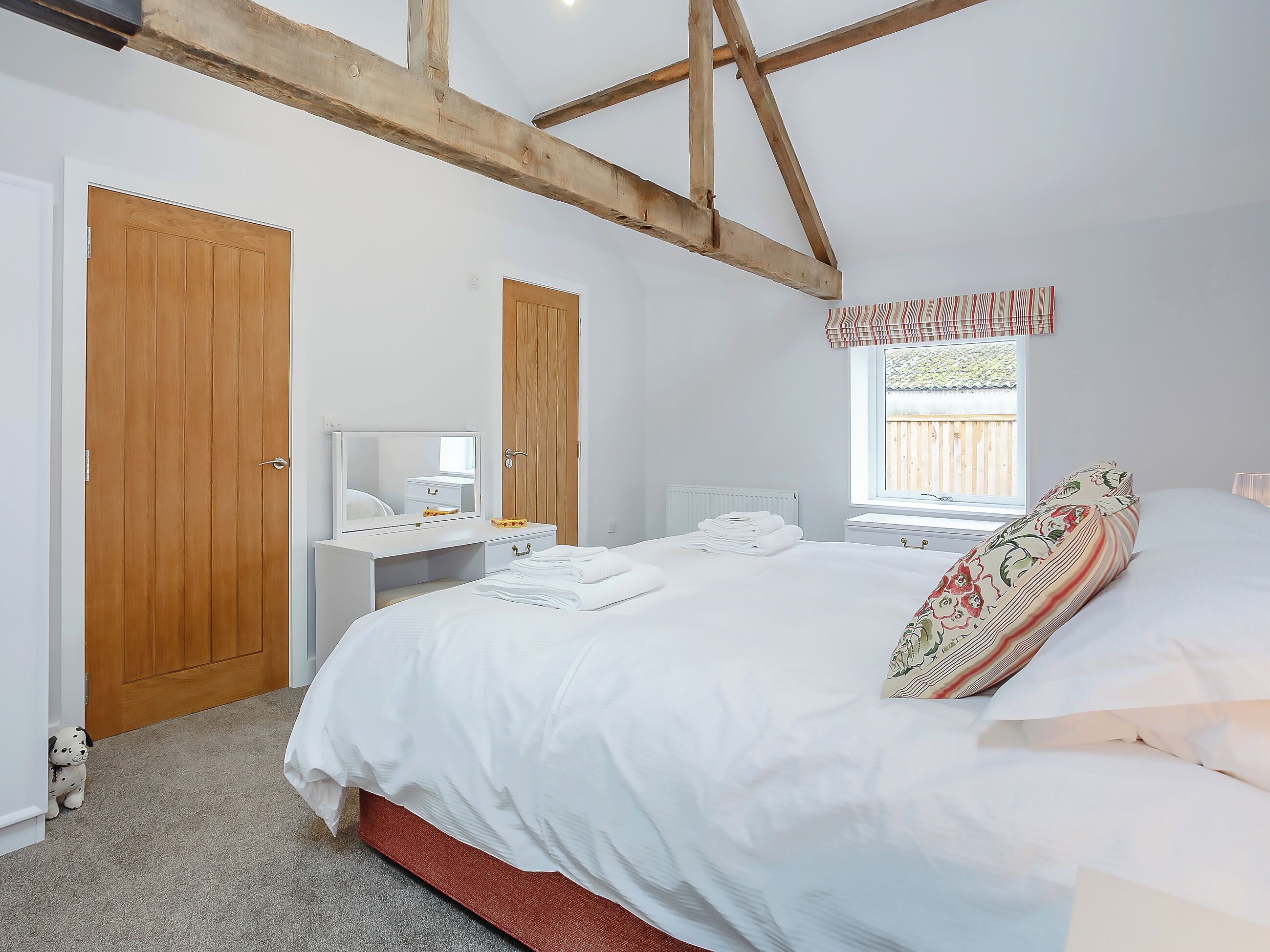 Een slaapkamer met een bed, een nachtkastje met spiegel, een raam met uitzicht op een houten schutting en een zichtbare houten balk aan het plafond.