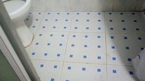 A bathroom with a toilet, white tiled floor with blue squares, and a visible drain. - Four Seasons Lodge (Naivasha)