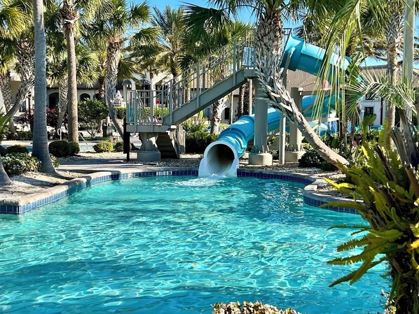 A pool with a blue slide, palm trees, and a building in the background.