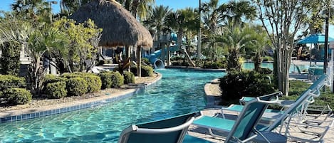 Un área de piscina con sillones reclinables, una estructura de paja, palmeras y un cielo azul despejado.