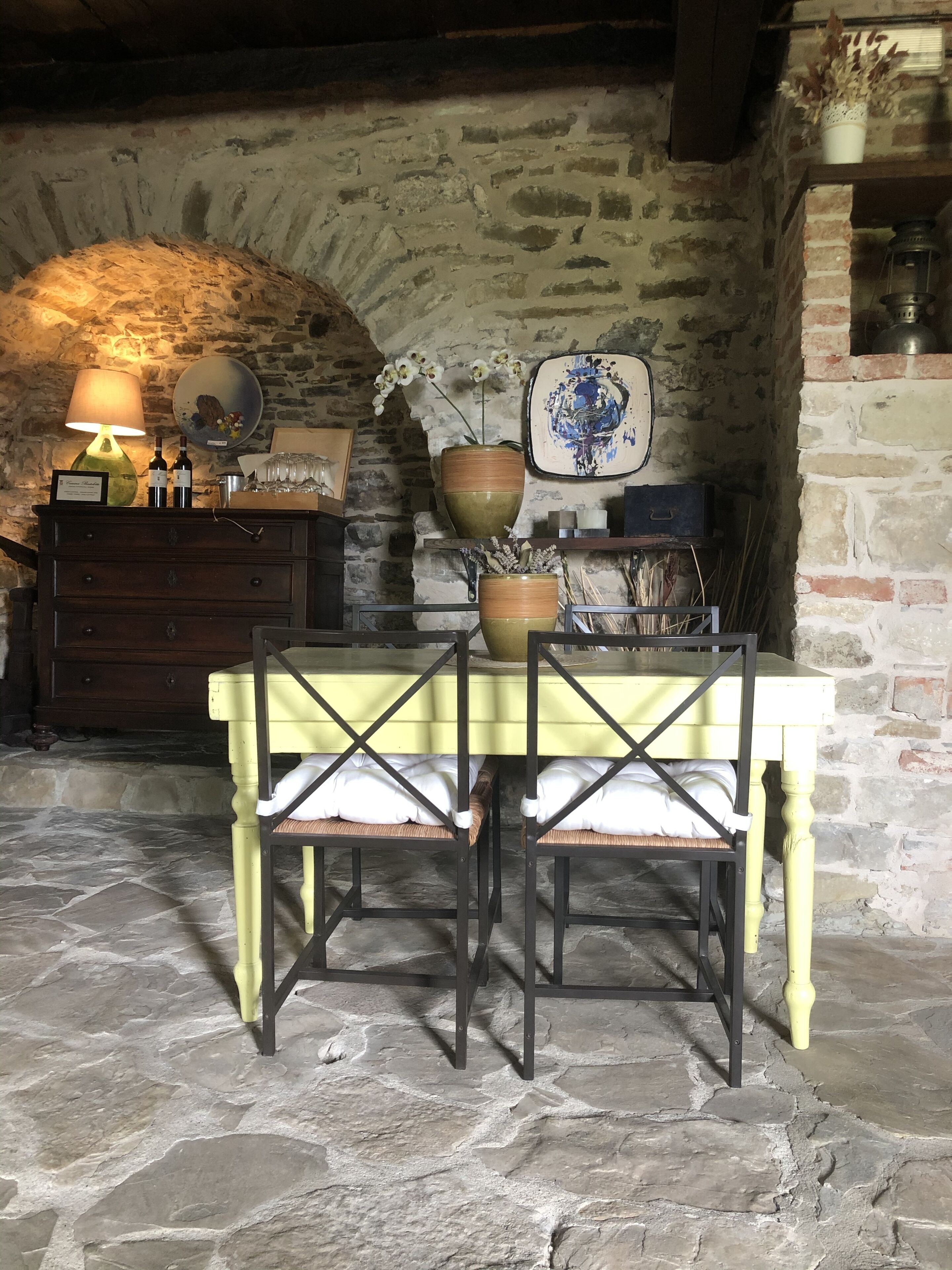 A rustic stone-walled room with a stone floor, a yellow table with two chairs, a wooden cabinet, and a wall-mounted shelf with decorative items.