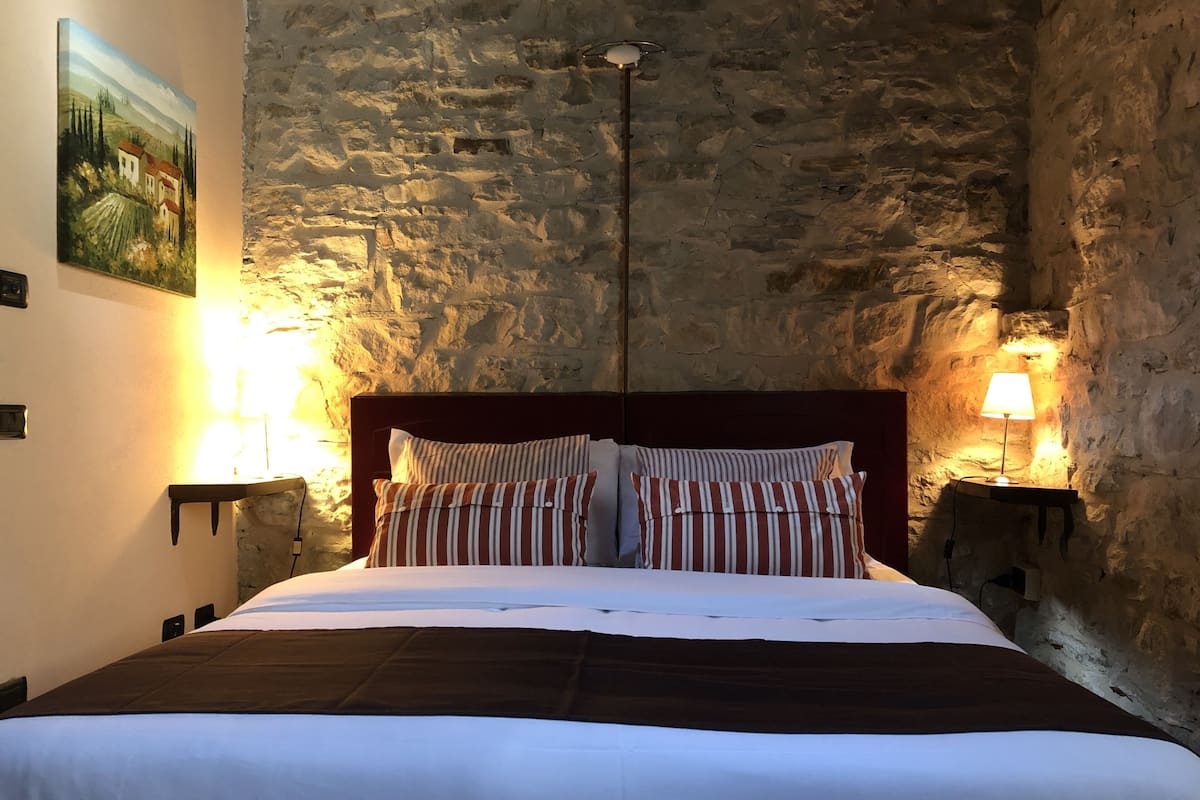 A bed in a room with stone walls, a painting on the wall, and two bedside lamps.