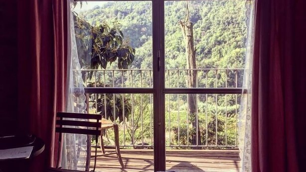 A room with a large arched window overlooking a lush forest. There is a bed with a striped blanket, a wooden chair, and a small table with a piece of paper on it. - Cascina Bertolotto Wine Resort. (Spigno Monferrato)