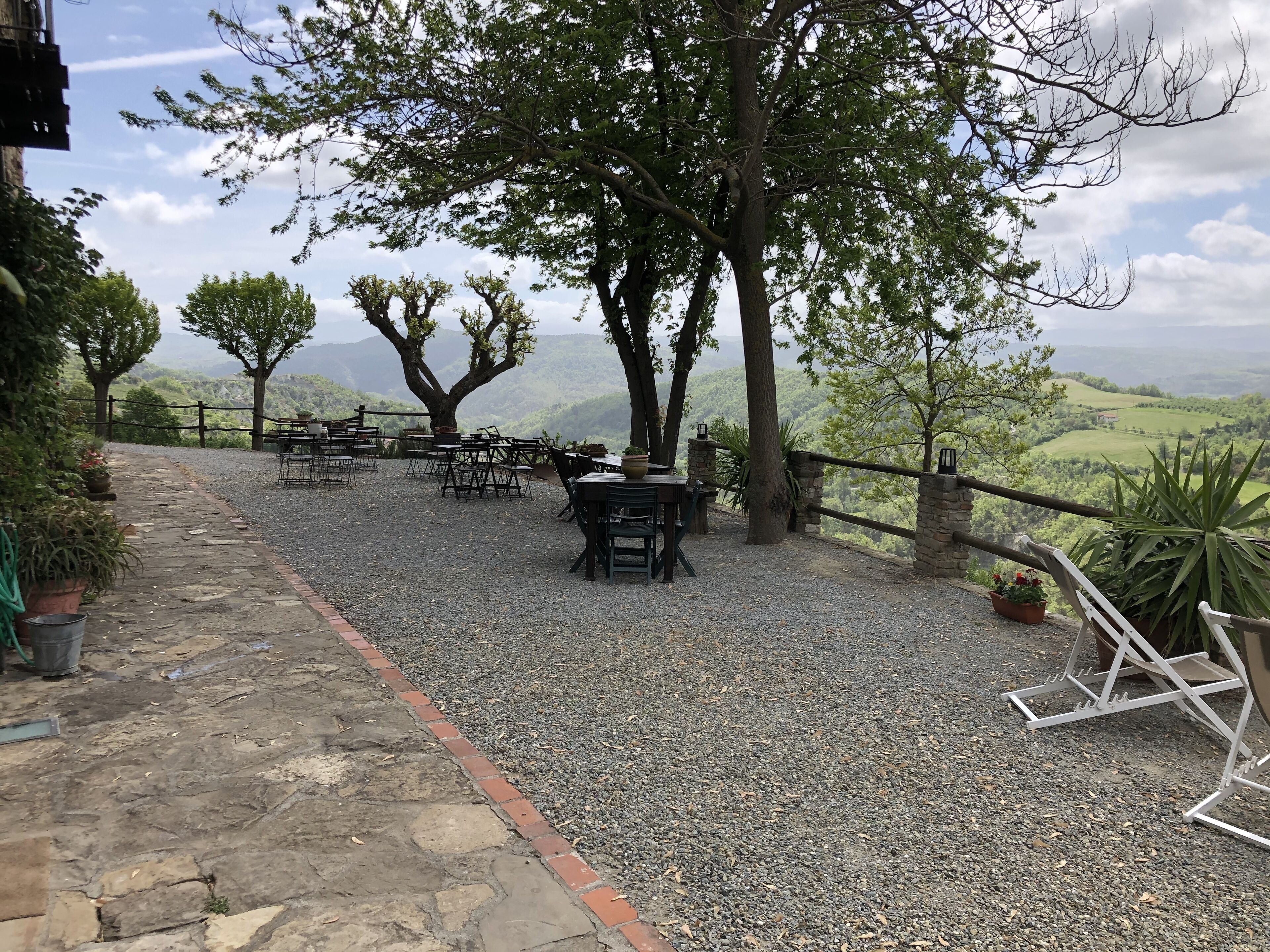 Un espacio al aire libre con mesas y sillas, rodeado de árboles y un muro de piedra, con vistas a un paisaje montañoso.