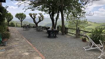 Un espacio al aire libre con mesas y sillas, rodeado de árboles y un muro de piedra, con vistas a un paisaje montañoso.