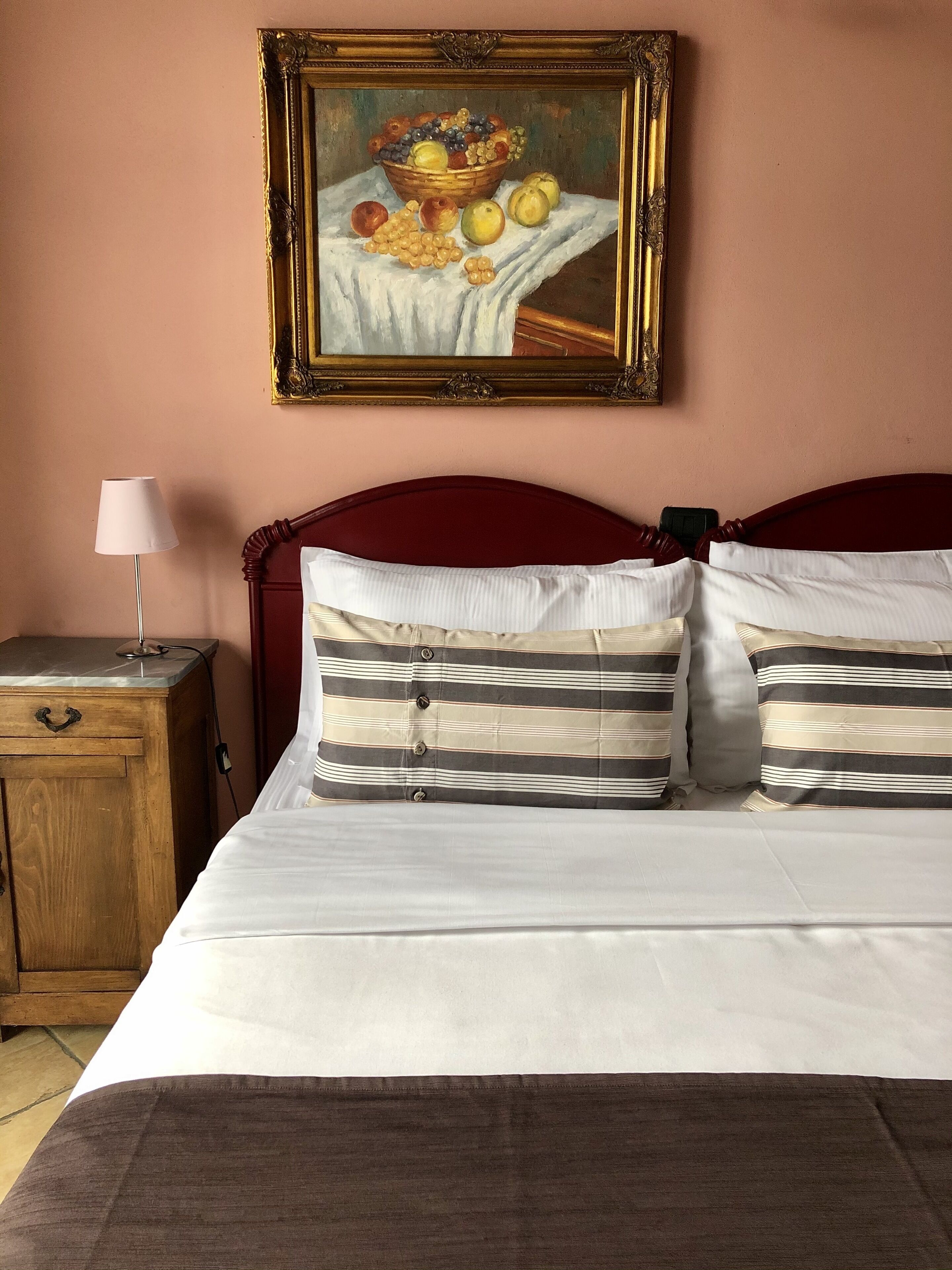 A hotel room with a bed, pillows, a nightstand, a lamp, and a painting of fruit on the wall.