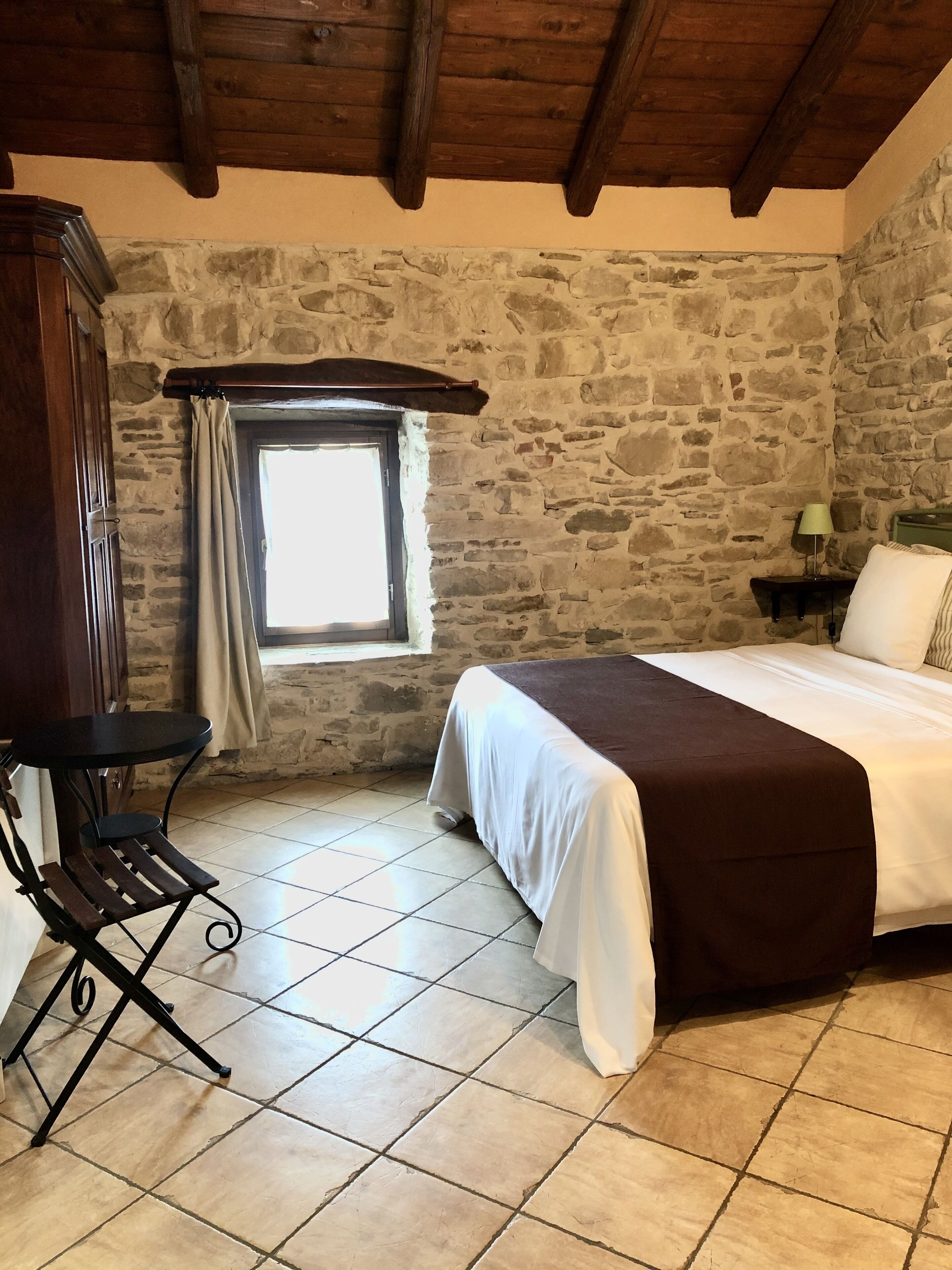 A bedroom with stone walls, a wooden ceiling, a bed with a white and brown bedspread, a small table with a lamp, and a chair.