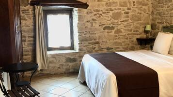 A bedroom with stone walls, a wooden ceiling, a bed with a white and brown bedspread, a small table with a lamp, and a chair.