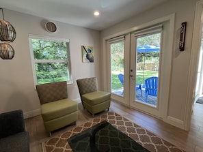 A living room with two green armchairs, a dark sofa, a patterned rug, a large window, and a sliding glass door leading to an outdoor patio with blue chairs and an umbrella.