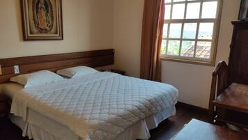 Uma cama bem arrumada com cobertura acolchoada, cabeceira de madeira, quadro religioso na parede e uma janela com cortinas.
