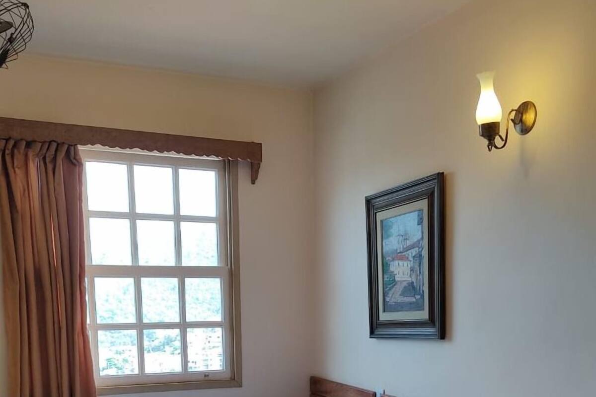 Ein Einzelbett mit Holz-Kopfteil, ein kleiner Nachttisch, eine Wandlampe und ein Fenster mit Vorhängen.