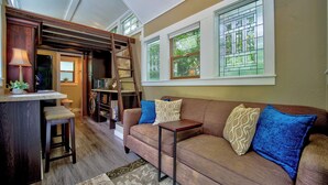 A cozy living area with a sofa, a small table, a ladder, and a window with a view of greenery. -  Tiny House Bungalow (Reynoldsburg)