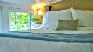 A bedroom with a bed, a nightstand with a lamp, and a view of greenery through a window. -  Tiny House Bungalow (Reynoldsburg)