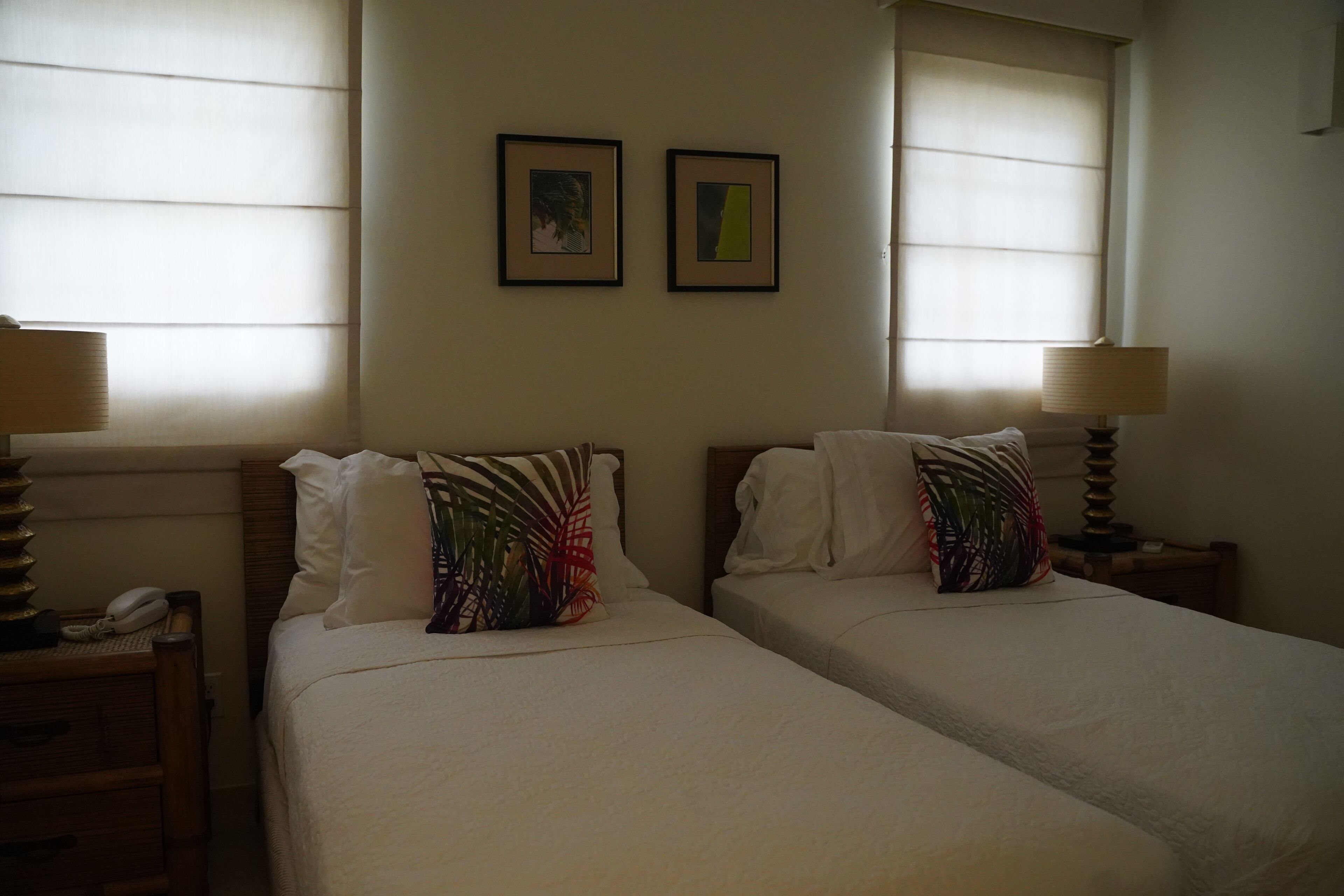 Habitación de hotel con dos camas, cada una con almohada y lámpara en la mesita de noche. Hay cuadros en la pared y una ventana con persianas.