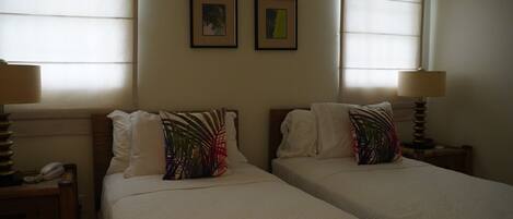Habitación de hotel con dos camas, cada una con almohada y lámpara en la mesita de noche. Hay cuadros en la pared y una ventana con persianas.