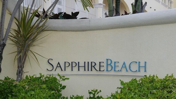 A sign for 'Sapphire Beach' with a white wall and green foliage in the foreground.