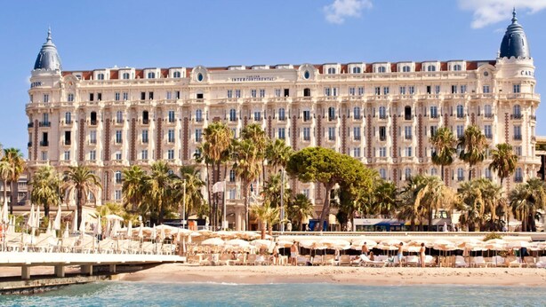 Et stort, historisk hotel med en imponerende facade, beliggende ved vandet, og et strandområde med liggestole og parasoller.