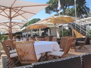 An outdoor dining area with wooden chairs and tables, umbrellas, and a staircase leading to an upper level. - 1BR CannesVacationRental by AmericanVacationLiving (Cannes)