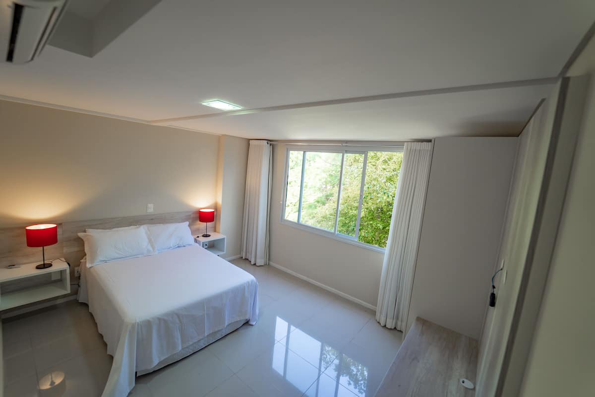 A hotel room with a large bed, two bedside tables with lamps, a window with curtains, and a view of greenery outside.