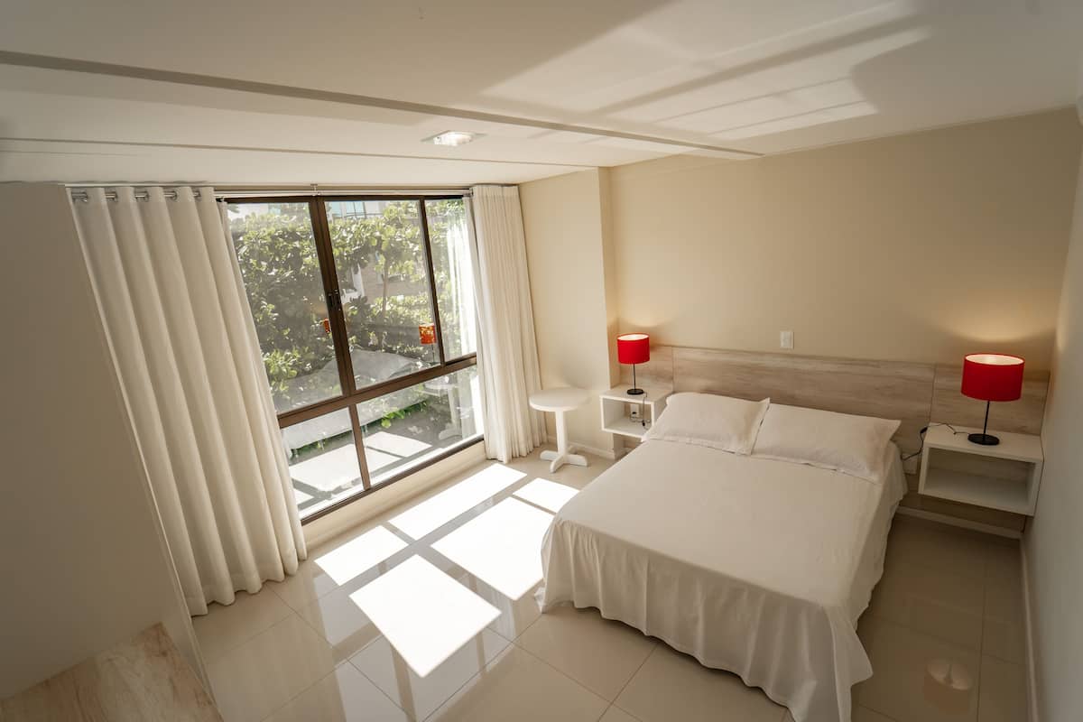 A modern hotel room with a large bed, two bedside tables with lamps, a small stool, and a large window with curtains.