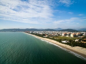 A coastal cityscape with a beach, residential buildings, and a hilly background. - Jurerê Sol & Mar Temporada (Florianópolis)