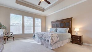 Een slaapkamer met een groot bed, een plafondventilator, een nachtkastje met een lamp en een dressoir met een bloemenstoel.