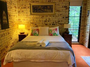 A room with a brick wall, a bed with floral pillows, two bedside lamps, and a window with a view.