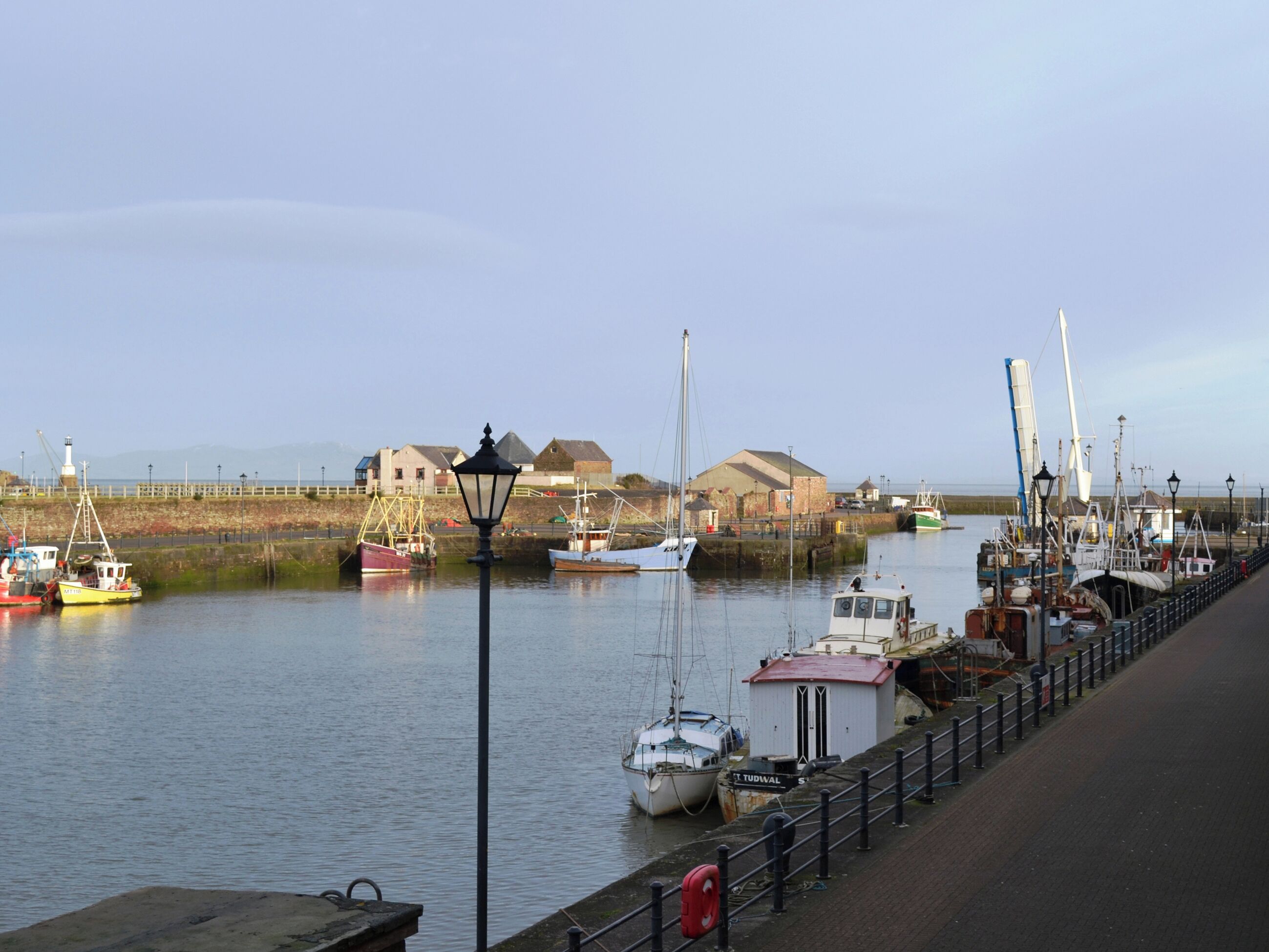 Un port où sont amarrés des bateaux, un phare et des bâtiments qui bordent la promenade.