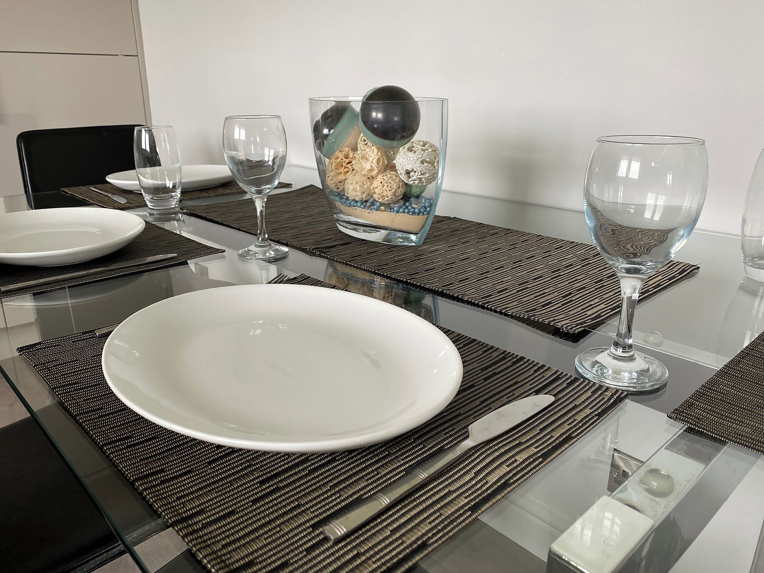 Een eettafel is gedekt met witte borden, bestek en glanzende wijnglazen. Op de tafel staat een decoratieve schaal met een zwarte bol en decoratieve objecten.
