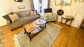 A living room with a grey sofa, a small armchair, a coffee table with a tray, a patterned rug, and a wall-mounted mirror.