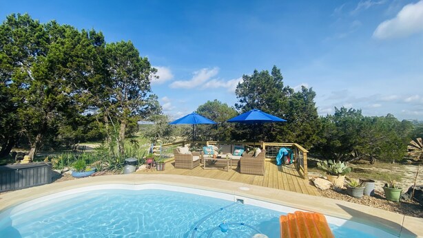 A swimming pool with a wooden deck, two blue umbrellas, and outdoor furniture.