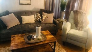 A living room with a dark grey sofa, a white armchair, a coffee table with remote controls, and a potted plant.