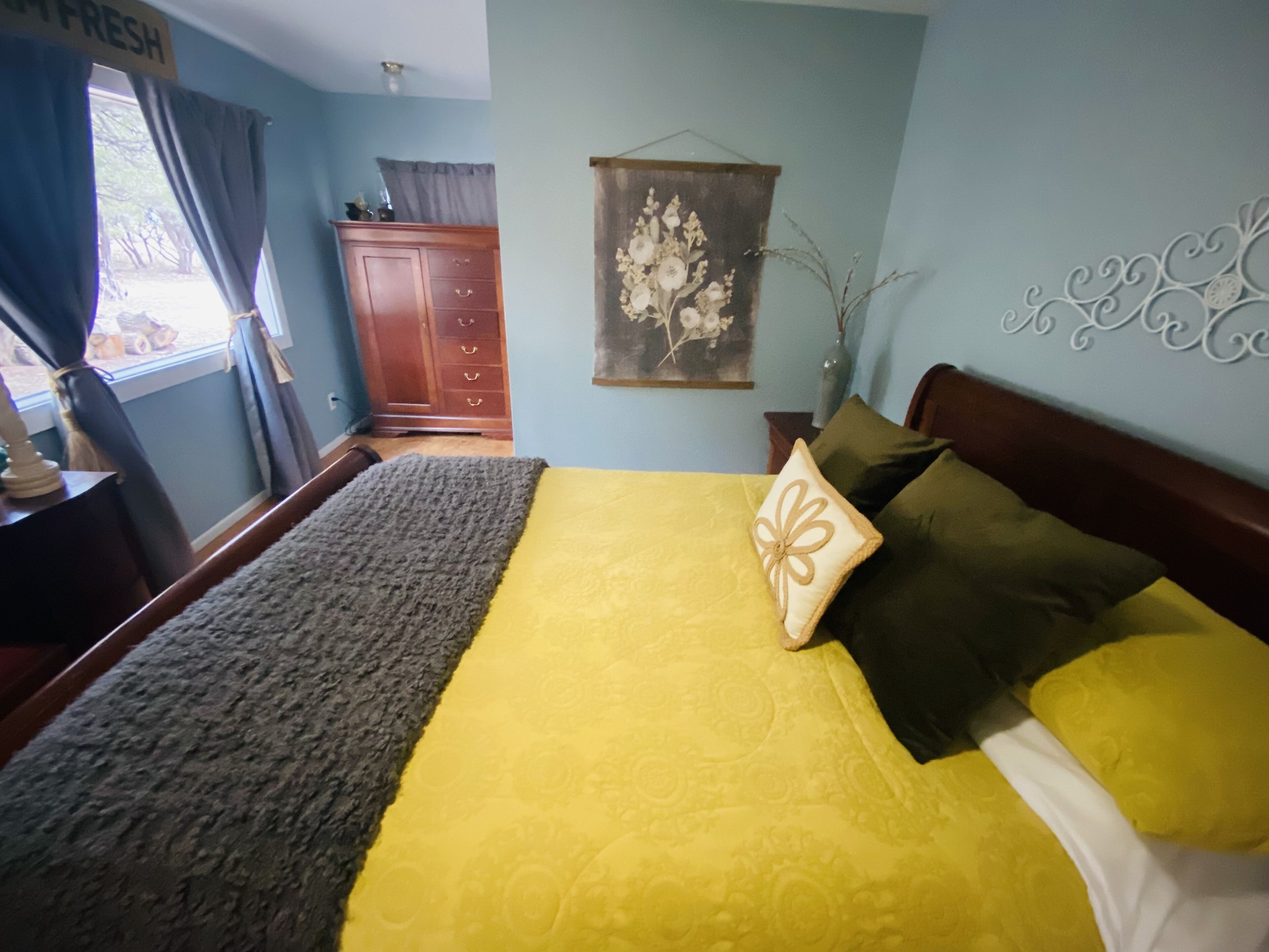 Un dormitorio con cama, cómoda, ventana con cortinas, un adorno de pared y un detalle decorativo.