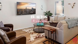 Un salon avec un tableau représentant un paysage urbain, un tapis en peau de vache, un canapé, une table basse et un crâne de vache accroché au mur.
