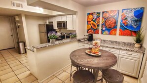Une cuisine compacte avec des meubles blancs, un plan de travail en granit, un réfrigérateur en acier inoxydable, un four à micro-ondes et une machine à café. Trois tableaux représentant des aliments aux couleurs vives ornent le mur.