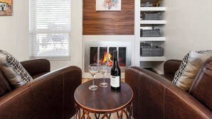 Un salon avec des canapés en cuir, une petite table en bois sur laquelle se trouvent une bouteille et des verres, une cheminée et une étagère ornée d’objets décoratifs.