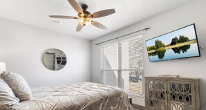 Un dormitorio con cama, ventilador de techo, televisión montada en la pared y ventana con persianas.