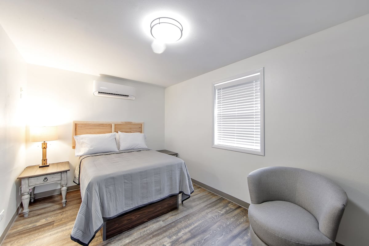 A bedroom with a bed, bedside table, lamp, air conditioning unit, window with blinds, and a chair.