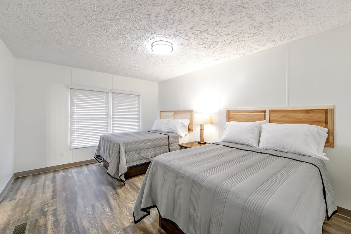 A bedroom with two beds, a wooden headboard, a bedside table with a lamp, and a window with blinds.