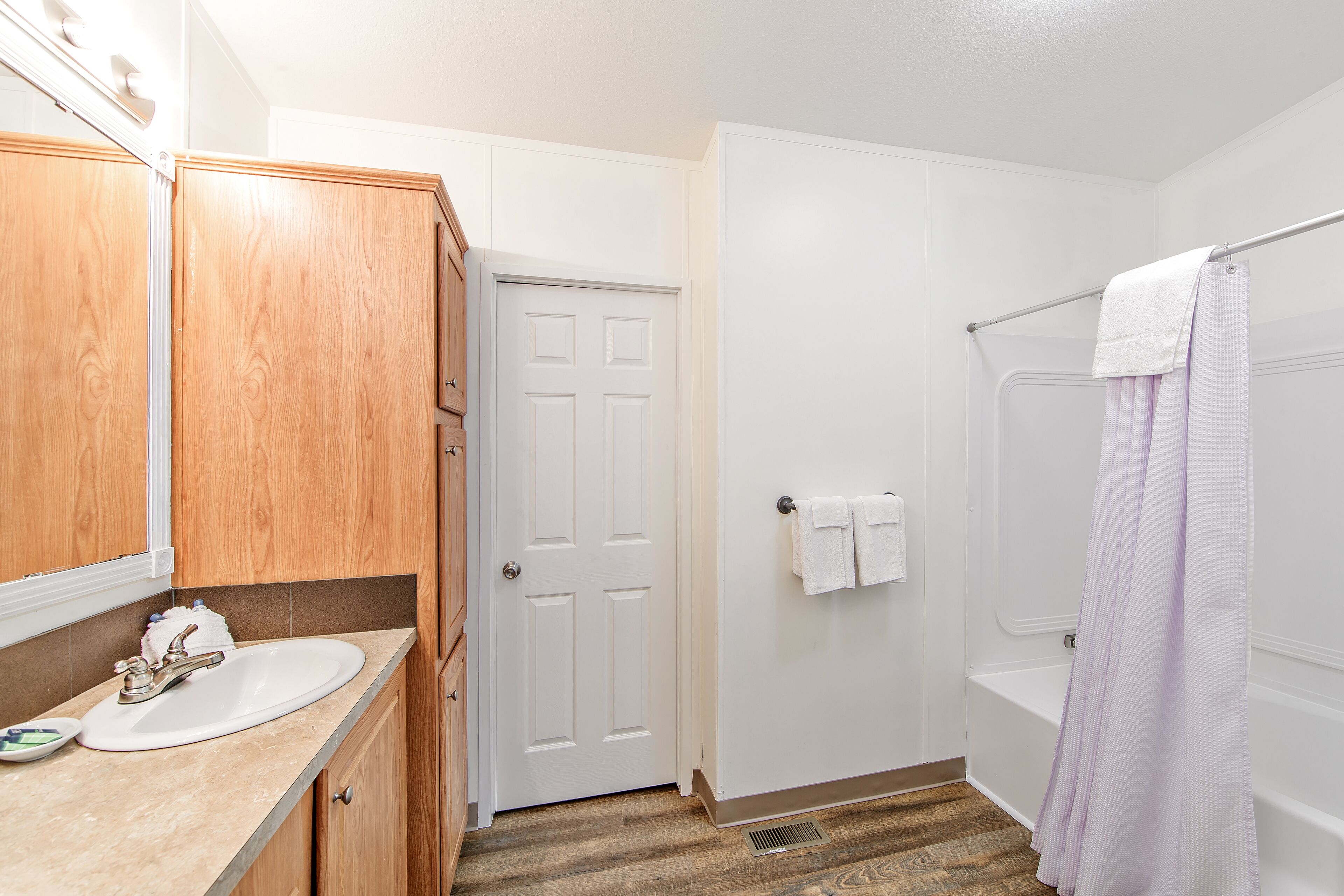 Une salle de bain avec un meuble-lavabo en bois, un lavabo blanc, une porte et une douche avec un rideau violet.