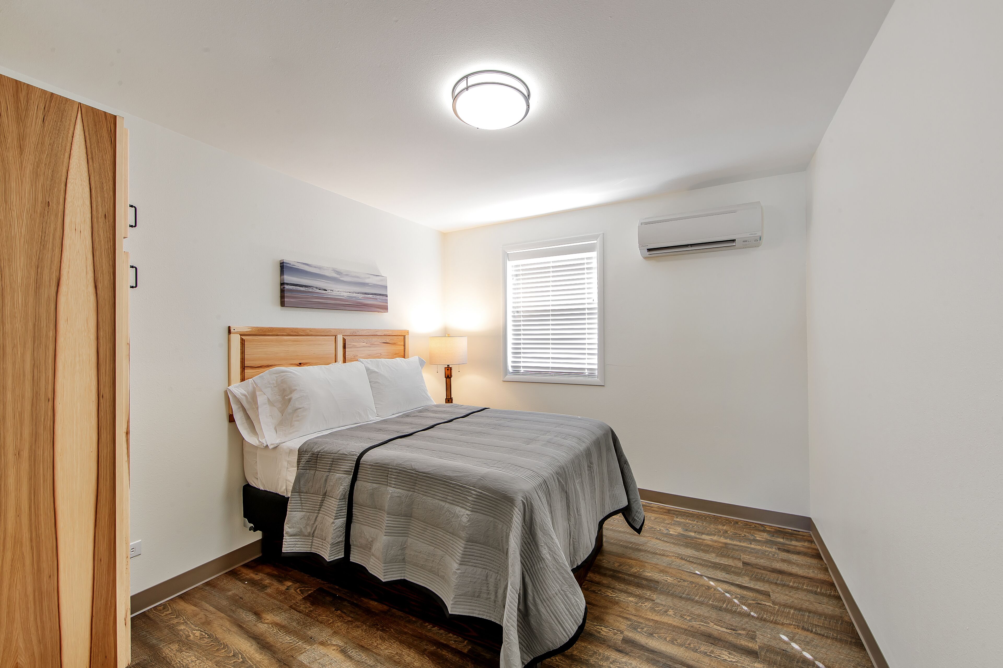 A bedroom with a wooden bed frame, a gray bedspread, a bedside lamp, a window with blinds, and an air conditioning unit on the wall.