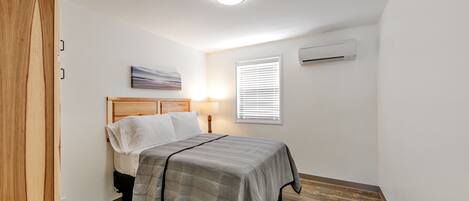 A bedroom with a wooden bed frame, a gray bedspread, a bedside lamp, a window with blinds, and an air conditioning unit on the wall.
